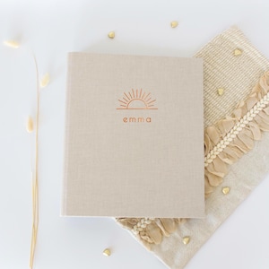 Può includere: Un diario beige con texture di lino con il nome "Emma" e un disegno a forma di sole in lamina di rame. Il diario è appoggiato su un panno decorativo beige con frange e coriandoli dorati a forma di cuore.