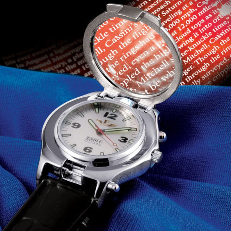 Puede incluir: Un reloj de pulsera plateado con una esfera blanca y una correa de cuero negra. El reloj tiene una lupa en la parte superior que se puede abrir para revelar una peque&ntilde;a ventana con texto. El texto dice "EAGLE OPTICAL".