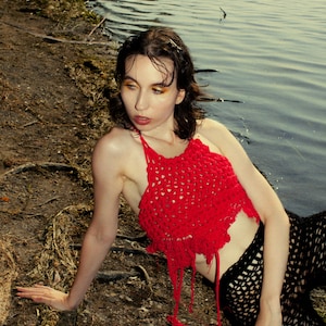 May include: A red crochet halter top with fringe detail. The top is paired with black, open-weave trousers. The person is near a body of water with a shoreline of dirt and roots.