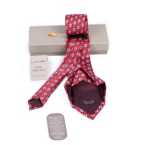 Op de afbeelding: Een rode zijden stropdas met een wit en blauw bloemenpatroon. De stropdas is gevouwen en wordt getoond op een doos met de merknaam "Seven ARM" en de tekst "%100 PURE SILK".