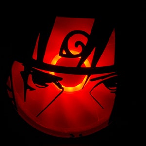 May include: A carved pumpkin with a silhouette of a character from the anime Naruto. The pumpkin is lit from inside, creating a glowing red effect.