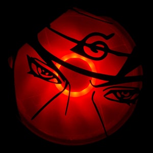 May include: A red, glowing, circular object with a black silhouette of a face with a headband and a symbol on it. The face has two eyes and a nose.