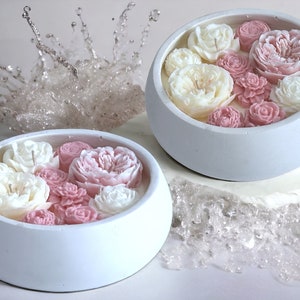 May include: Two white ceramic bowls filled with white and pink wax flowers. The bowls are on a white surface with a clear, sparkly background.