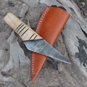 Könnte beinhalten: Ein kleines, handgefertigtes Messer mit einer gemusterten Klinge aus Stahl und einem hellbraunen Holzgriff. Das Messer befindet sich in einer braunen Lederscheide.