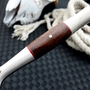 May include: Close-up of a knife handle with a dark wood grip, white sections and a thin red band. The handle has a brass rivet and a patterned blade. A rope and a decorative object are in the background.