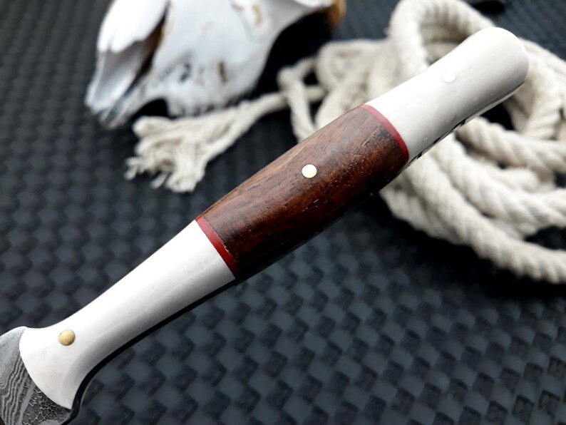 May include: Close-up of a knife handle with a dark wood grip, white sections and a thin red band. The handle has a brass rivet and a patterned blade. A rope and a decorative object are in the background.