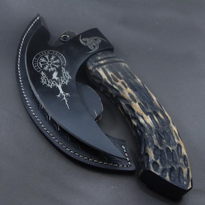 Puede incluir: Un hacha negra con una funda de cuero y un mango con un patrón marrón y negro. La cabeza del hacha presenta un diseño de inspiración vikinga con una brújula, un árbol, un pájaro y un nudo.