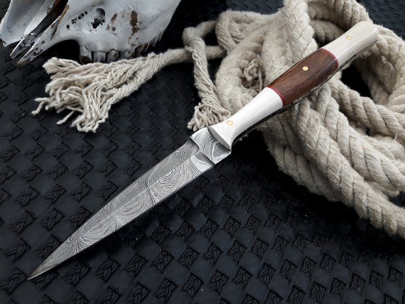 May include: A Damascus steel dagger with a patterned blade and a handle made of wood and bone. The handle has a dark wood centre with brass pins and a red accent. A coiled rope and a skull are in the background.