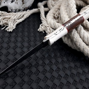 May include: A handmade knife with a dark blade and a handle combining light and dark wood, with red and white accents. The knife is shown on a textured black surface with a coiled rope in the background.