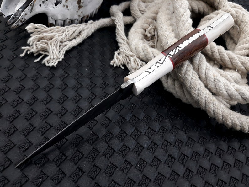 May include: A handmade knife with a dark blade and a handle combining light and dark wood, with red and white accents. The knife is shown on a textured black surface with a coiled rope in the background.