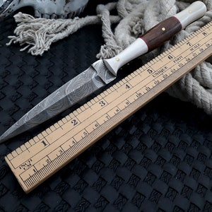 May include: A handcrafted dagger with a patterned blade and a wooden handle. The handle features a white section and is accented with brass pins. A wooden ruler, marked in inches, is placed alongside the knife, indicating its size.