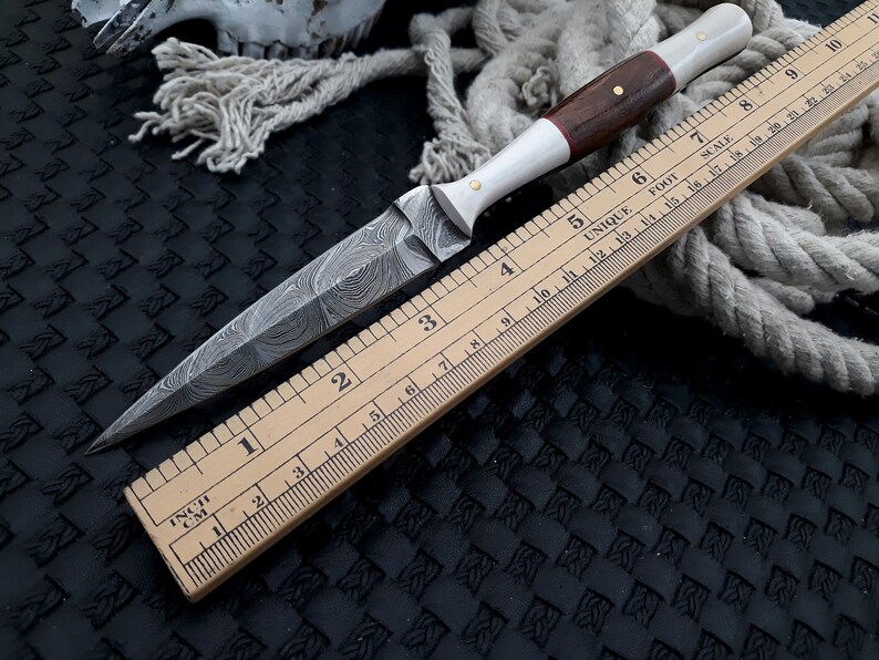 May include: A handcrafted dagger with a patterned blade and a wooden handle. The handle features a white section and is accented with brass pins. A wooden ruler, marked in inches, is placed alongside the knife, indicating its size.
