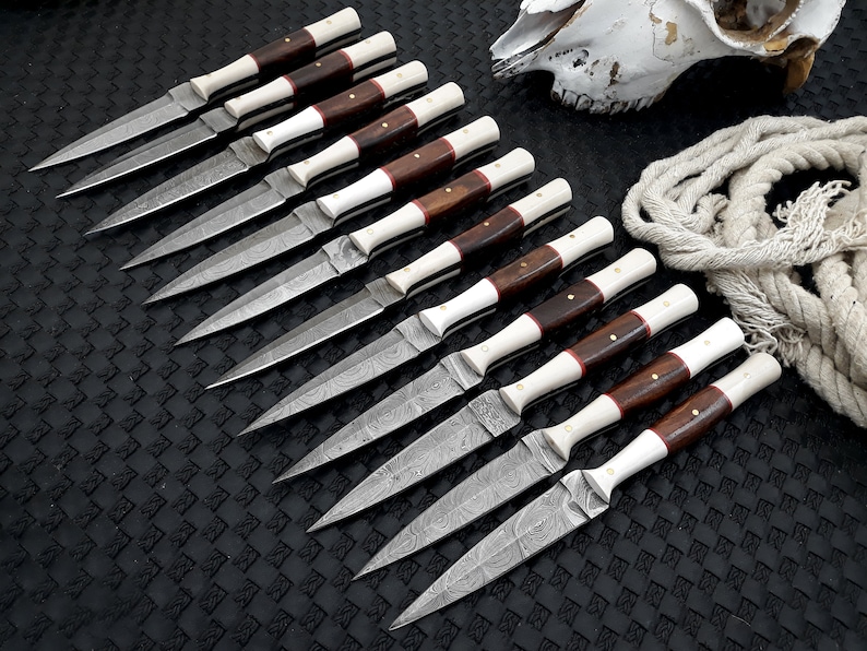 May include: Ten knives with patterned steel blades, dark wood handles, and white and red accents. The knives are arranged on a black surface, with a skull and rope in the background.