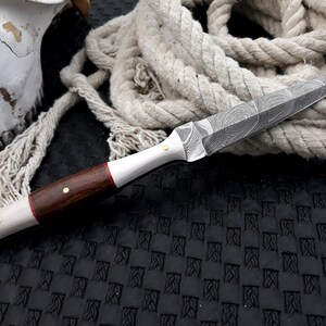 May include: A Damascus steel knife with a wooden handle. The blade features a distinctive swirling pattern. The handle combines dark wood, bone, and brass rivets. A rope and skull are in the background.
