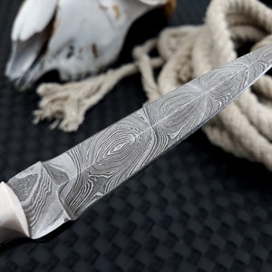 May include: Close-up of a Damascus steel knife with a cream-coloured handle and a brass rivet. The blade features a distinctive swirling pattern. A coiled rope and a skull are in the background.