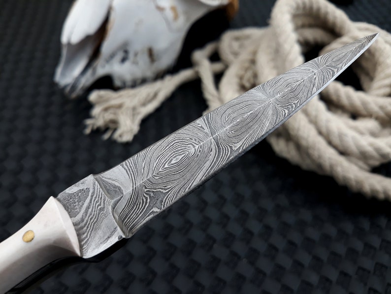 May include: Close-up of a Damascus steel knife with a cream-coloured handle and a brass rivet. The blade features a distinctive swirling pattern. A coiled rope and a skull are in the background.