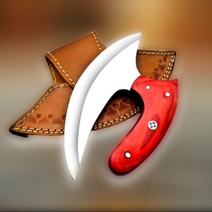 Op de afbeelding: Een gebogen mes van roestvrij staal met een rode houten handgreep en een leren schede. Het mes heeft een uniek, gebogen blad en een comfortabele handgreep.