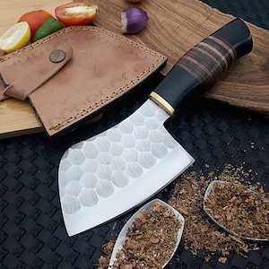May include: A cleaver knife with a textured blade and a dark handle with wood accents. The knife has a leather sheath and is displayed with fresh vegetables and spices, suggesting culinary use.
