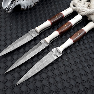 May include: Three Damascus steel knives with ornate blades and handles. The handles feature dark wood, bone-coloured material, and red accents. The knives are arranged on a textured black surface.