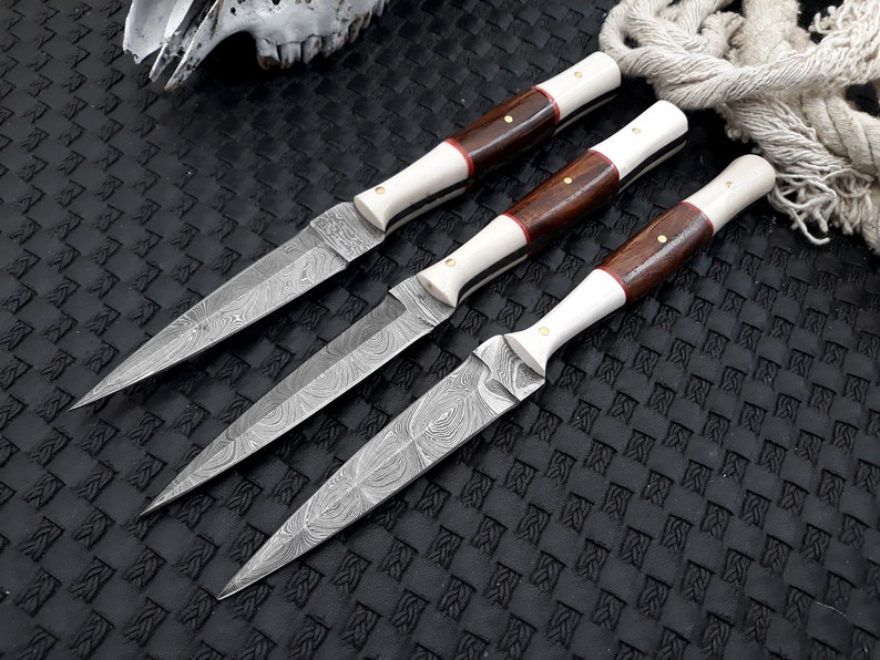 May include: Three Damascus steel knives with ornate blades and handles. The handles feature dark wood, bone-coloured material, and red accents. The knives are arranged on a textured black surface.