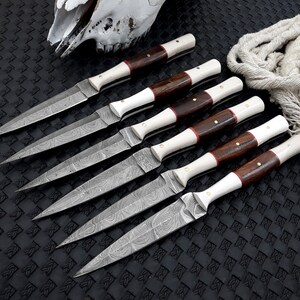 May include: Six handmade knives with patterned blades, wooden handles, and white accents are arranged on a textured black surface. The knives have a sharp, pointed design. A skull and rope are in the background.