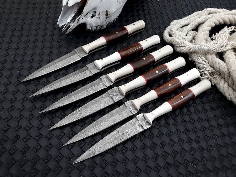 May include: Six handmade knives with patterned blades, wooden handles, and white accents are arranged on a textured black surface. The knives have a sharp, pointed design. A skull and rope are in the background.