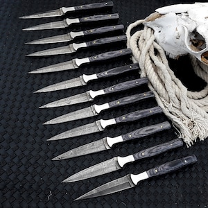 May include: A set of 12 Damascus steel knives with black wood handles. The knives are arranged in a row on a black surface.