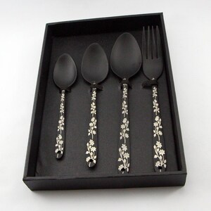 May include: A set of four black metal spoons and a fork with a floral design. The utensils are in a black box.