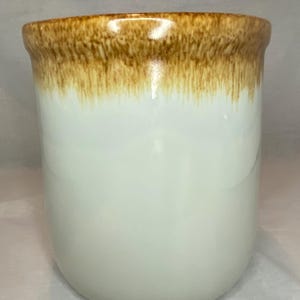 May include: A white ceramic mug with a brown, dripped glaze around the rim. The mug has a smooth, glossy finish and a rounded bottom. The mug is a simple, yet elegant design.