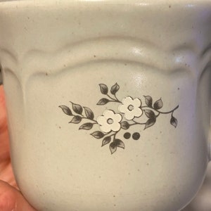 May include: A white ceramic mug with a floral design in brown and white. The design features a branch with leaves and two small flowers.