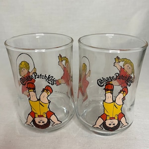 May include: Two clear glass drinking glasses with a Cabbage Patch Kids design. The glasses feature a cartoon image of a Cabbage Patch Kid with blonde hair, wearing a red and yellow outfit, and holding a jump rope. The text "Cabbage Patch Kids" is printed on the glass in a black and red font.