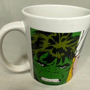 May include: White ceramic mug featuring a comic book illustration of the Hulk's face in green, black, and white. The mug has a curved handle and a glossy finish. The design is a bold, colorful graphic.