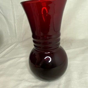May include: A ruby red glass vase with a bulbous base and a flared top. The vase has a series of horizontal ridges near the top of the base. The glass is smooth and reflective, with a glossy finish.