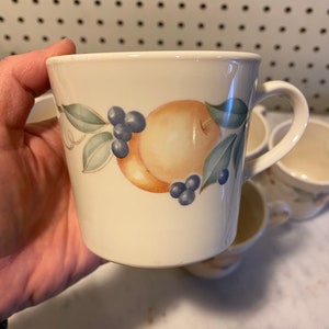 May include: A white ceramic mug with a fruit design. The design features a peach, leaves, and blueberries. The mug has a handle.