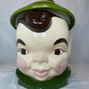 May include: A ceramic cookie jar shaped like a head with a glossy finish. The figure has a cream-colored face, dark brown hair, pointed ears, and pink cheeks. It wears a green hat and a matching green collar, featuring a smiling expression.