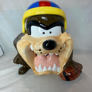 May include: A ceramic cookie jar shaped like the Tasmanian Devil character wearing a yellow, blue, and red football helmet. The figure has brown fur, a wide open mouth with white teeth, and is holding a brown football in one of its paws.