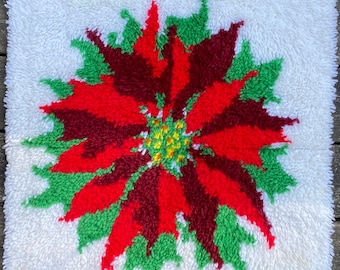 May include: A square, white, tufted wall hanging featuring a poinsettia design. The flower has red and burgundy petals with green leaves and a yellow center. Gold-colored hooks are attached to the top edge for hanging.