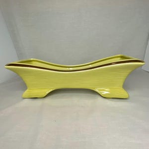 May include: A light yellow ceramic planter with a brown accent line. The planter has a curved, elongated shape with a ribbed texture and a mid-century modern design. It features a wide opening and is supported by two flared feet.
