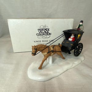 May include: A porcelain figurine of a brown horse pulling a black carriage with a driver and passenger, set on a white snow-like base. Behind it sits a white box labeled The Heritage Village Collection Kings Road Cab Handpainted Porcelain Accessory.