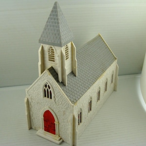 May include: A miniature church model with a gray roof and steeple, and a red door. The building is off-white with arched windows and a textured facade. The model is a detailed representation of a church.