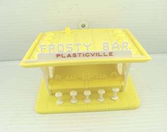 May include: A yellow plastic toy bar with the words "Frosty Bar" and "Plasticville" in red and white lettering. The bar has a roof, a counter, and five white stools. The toy is a miniature replica of a bar.