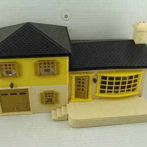 May include: A yellow and beige toy house with a black roof, garage, and chimney. The house has multiple windows with yellow frames and shutters. The front door is open, and there is a small porch.