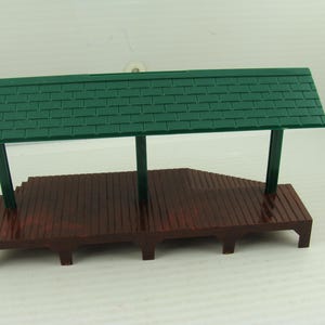 May include: A miniature train station platform model featuring a dark brown wooden-style base with vertical plank detailing. The structure is supported by three dark green pillars that hold up a rectangular green roof with a shingle pattern.