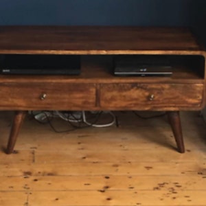 May include: A wooden TV stand in a mid-century modern design. It has two drawers with brass handles, a shelf, and tapered legs. Two black electronic devices sit on the shelf. The stand is approximately 122 cm wide.