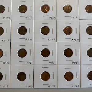 May include: A collection of copper-colored Lincoln pennies from the 1970s, each encased in a white cardboard holder. The dates and mint marks are visible, including 1970-D, 1971-D, 1972-D, 1973, 1975-S, 1970-S, 1971-S, 1972-S, 1973-D, 1974, 1974-D, 1976, 1977, 1978, 1979, 1975, 1976-D, 1977-D, 1978-D, and 1979-D.