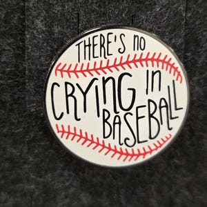 May include: A circular pin badge with a baseball design. The white badge has red stitching and the words "THERE'S NO CRYING IN BASEBALL" in black. The background is a dark grey fabric, providing a contrasting backdrop for the pin.