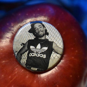 May include: A red apple with a button pinned on top. The button features a black and white image of a man wearing an Adidas tank top and a backwards cap. The background is a blurred blue.