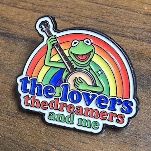 Könnte beinhalten: Ein Emaille-Anstecker aus Metall, der einen grünen Frosch zeigt, der vor einem bunten Regenbogen Banjo spielt. Der Text "the lovers the dreamers and me" ist in blauen, roten und grünen Buchstaben am unteren Rand des runden Designs abgebildet.