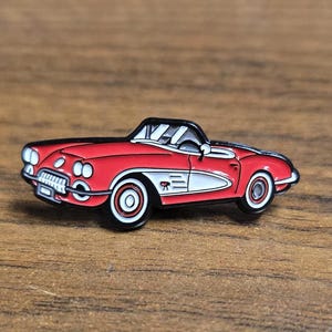 Peut inclure: Une épingle de voiture décapotable rouge vintage avec des accents blancs. L'épingle présente un toit noir, des roues à jantes blanches et une calandre avant détaillée. La voiture est posée sur un fond en bois.