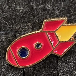 Puede incluir: Un pin de esmalte rojo y amarillo con forma de cohete y contorno dorado. El pin presenta un círculo azul en la parte delantera y un círculo azul más pequeño cerca del centro. Las aletas del cohete son amarillas.